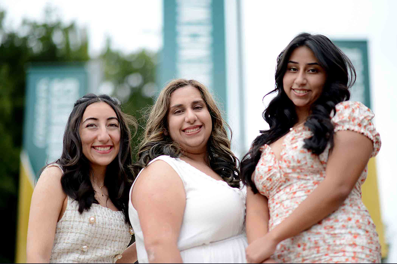 Three young women in white dresses smile for a portrait in front of Cal Poly banners.