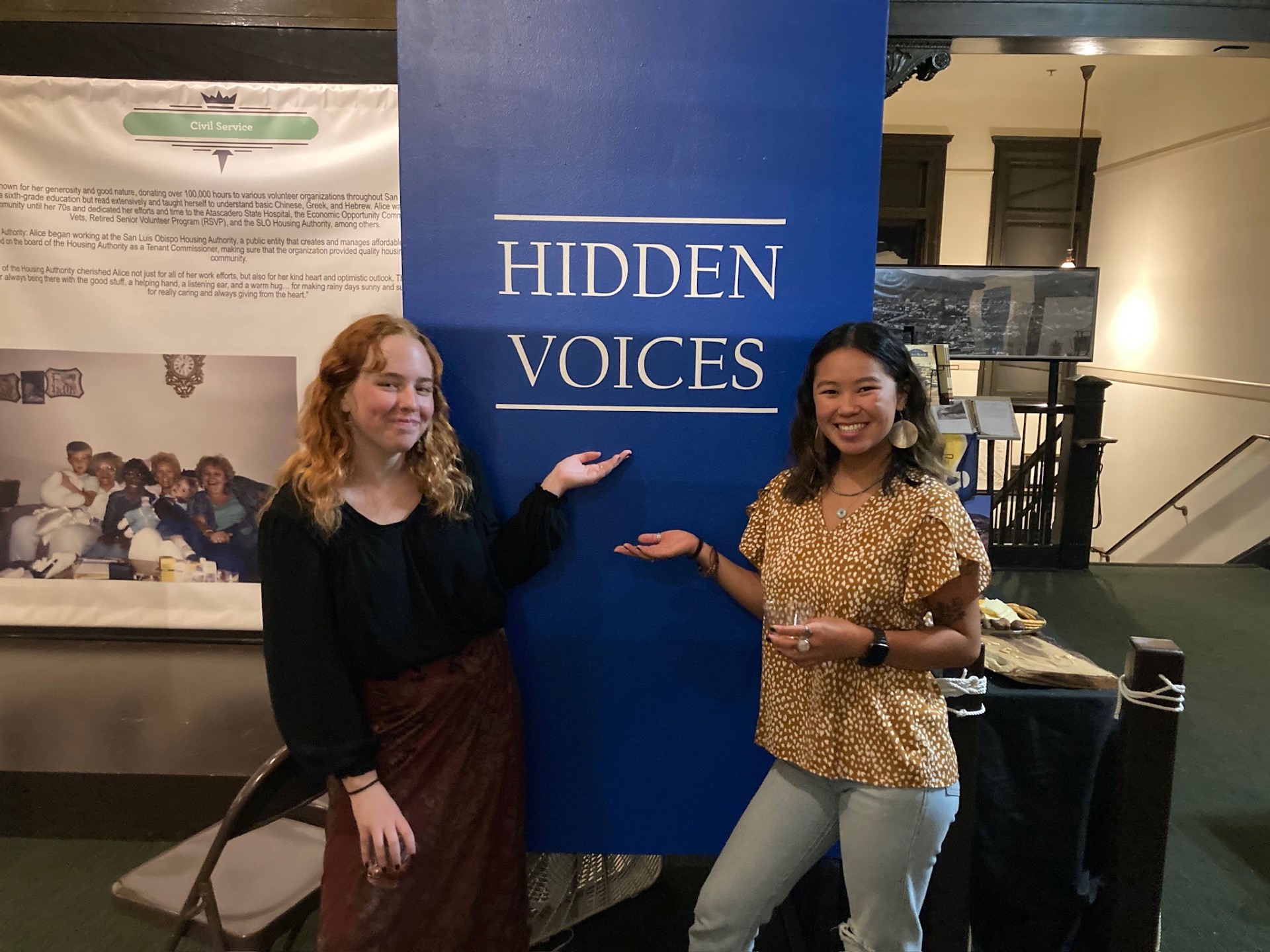 Two students stand in front of a blue poster reading 'Hidden Voices' during opening night of a historical exhibit.